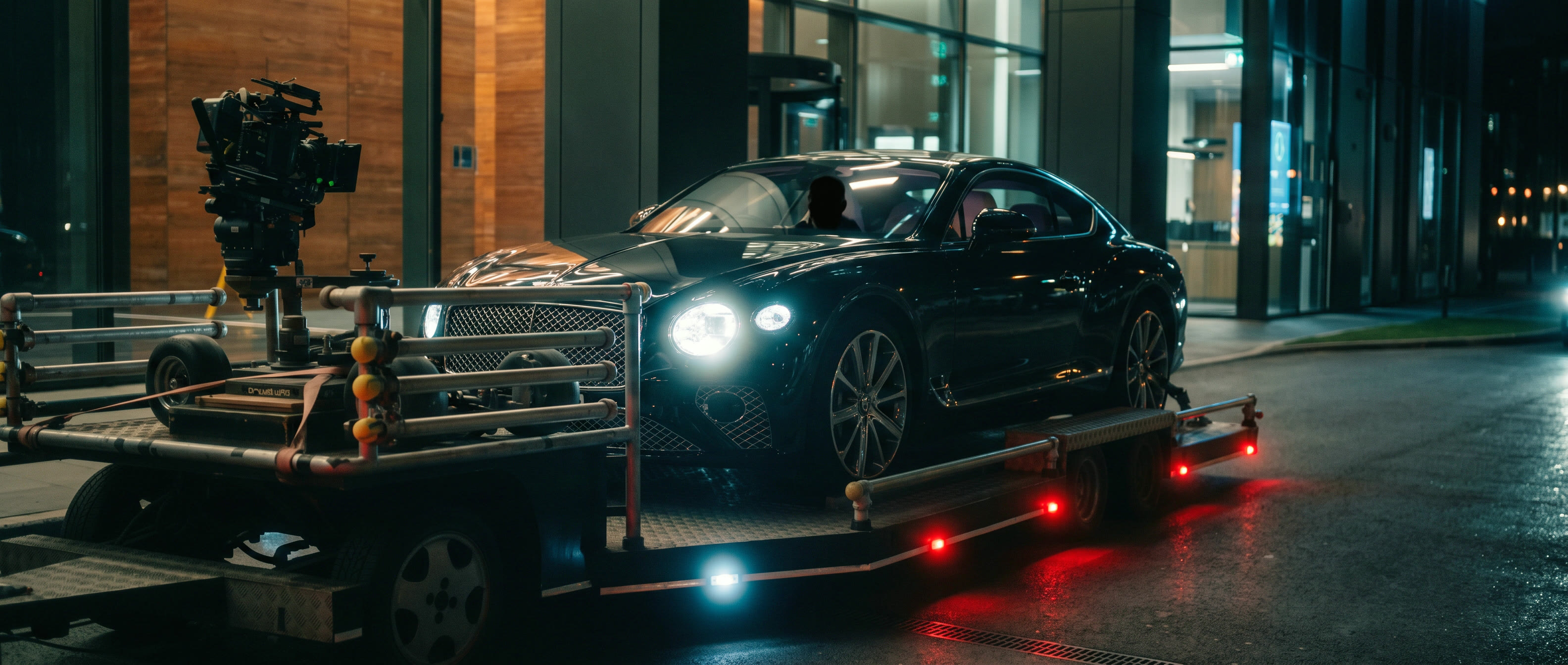 A black Bentley on a Cinema Cars low loader at night, a cinema camera rigged on the deck in front of it